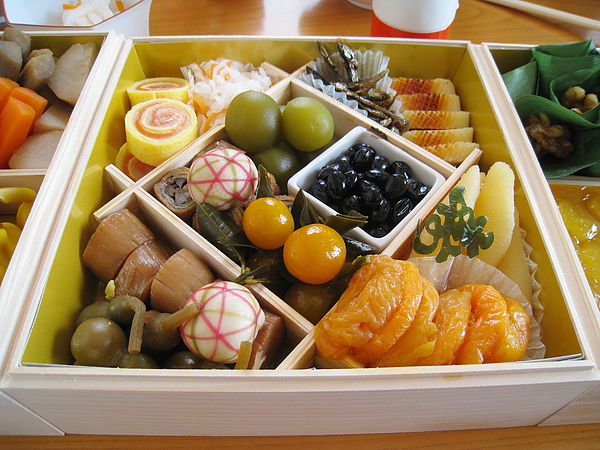 第二層中央菜色：右邊瓷盒內是甜黑豆、下方是金蜜柑煮、上方是梅甘露煮、左方底部是穴子八幡卷、彩線小球是手毬麩