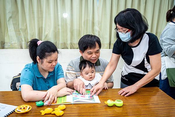 （圖說：新北市立圖書館閱讀推廣志工（圖右）協助視障家長帶孩子一起跟著講師指示觸摸書籍。）