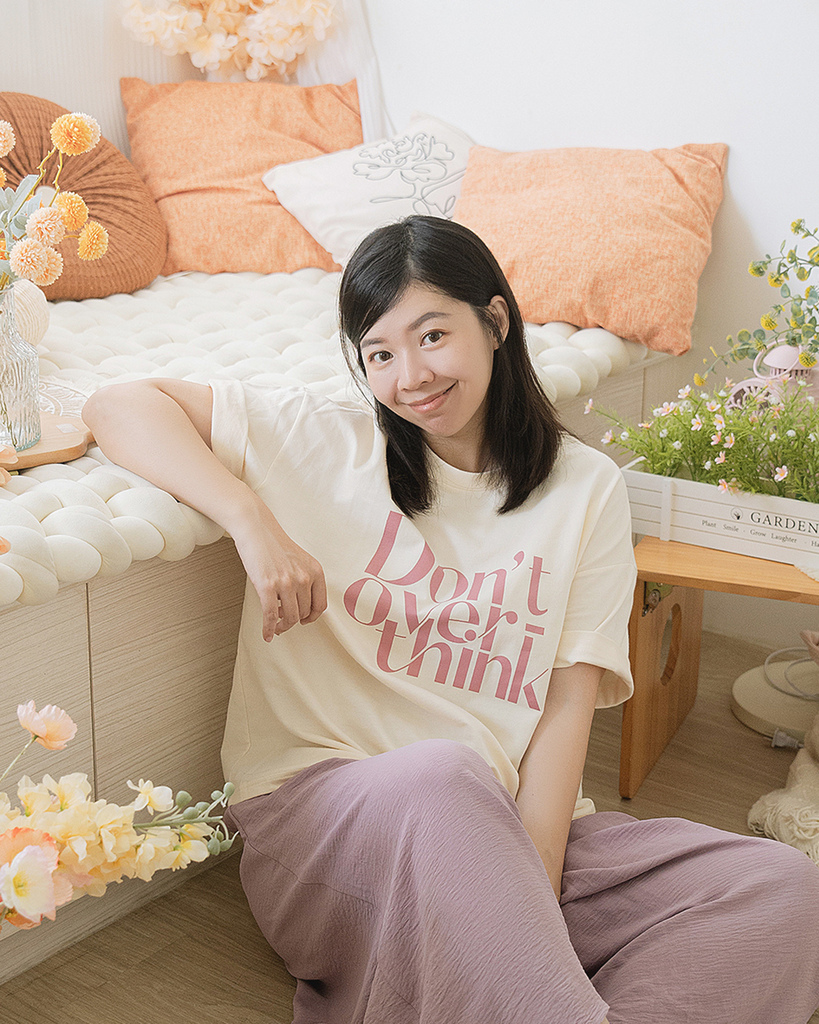 大尺碼女孩穿著白色印花T恤與紫色寬褲，站在簡約溫馨的空間中，搭配植物與自然元素，展現2025夏季穿搭的舒適度假感與粉嫩穿搭風格