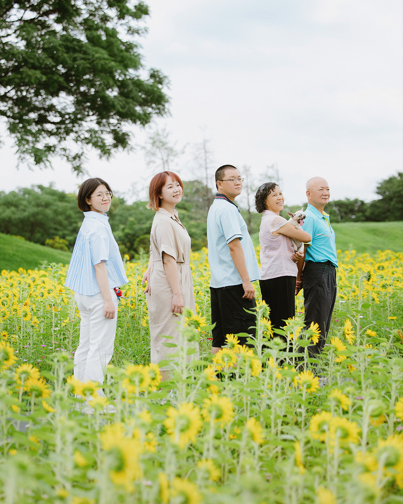 台中向日葵花海寵物全家福母親節自然風格寵物寫真紀錄 五位家人站在台中向日葵花田中合影,背景是綠樹與雲層,畫面自然溫馨。此照片為母親節寵物寫真的一部分,展現高齡狗狗與家人間的真實陪伴與情感連結,屬於自然風格的寵物全家福拍攝
