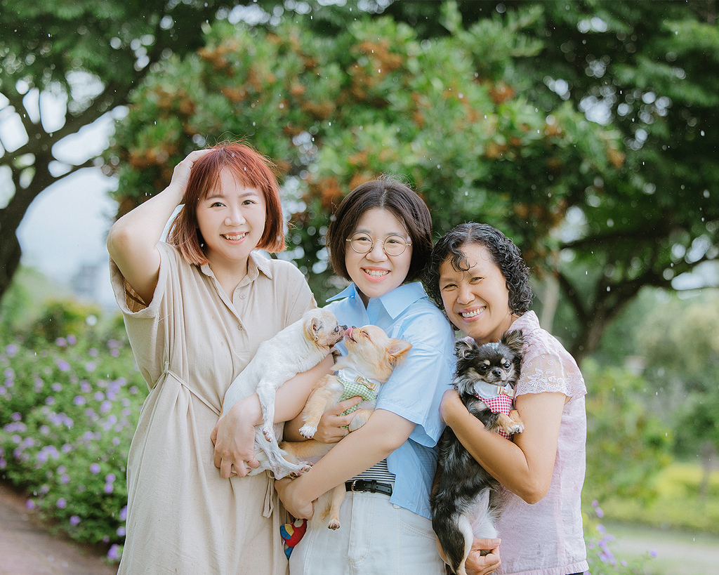 母親節寵物寫真台中花園全家福 高齡吉娃娃與家人的自然合影 三位女性在台中花園中合影,其中兩人分別抱著一隻高齡吉娃娃與穿著蝴蝶結的黑色小型犬。背景是綠樹與花卉,畫面溫馨自然,展現寵物全家福寫真的真實陪伴與情感連結。