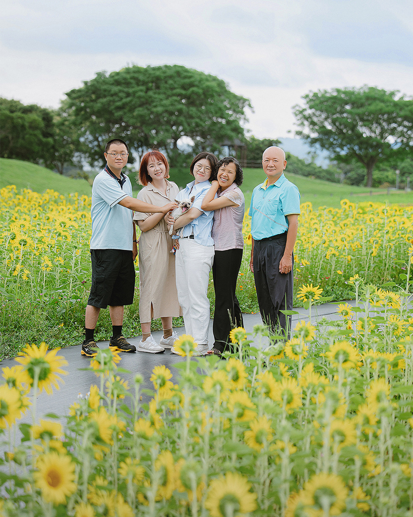 台中向日葵花海寵物全家福母親節自然風格寵物寫真推薦 五位家人站在台中向日葵花田中的步道上合影,背景是綠樹與雲層,畫面自然溫馨。此照片為母親節寵物寫真的一部分,展現毛孩與家人間的真實陪伴與情感連結,屬於自然風格的寵物全家福拍攝