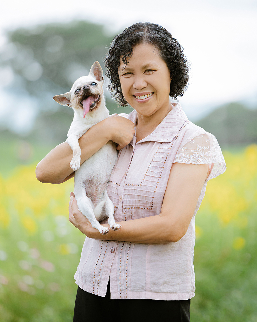 台中自然風格寵物寫真母親節全家福 高齡狗狗花園合影紀錄 一位穿著粉色蕾絲袖上衣的女性在台中花園中抱著一隻高齡白色吉娃娃,狗狗神情愉悅、舌頭微吐,背景為綠葉與黃色花卉。此照片為母親節寵物寫真的一部分,展現毛孩與主人間的真實陪伴與情感連結